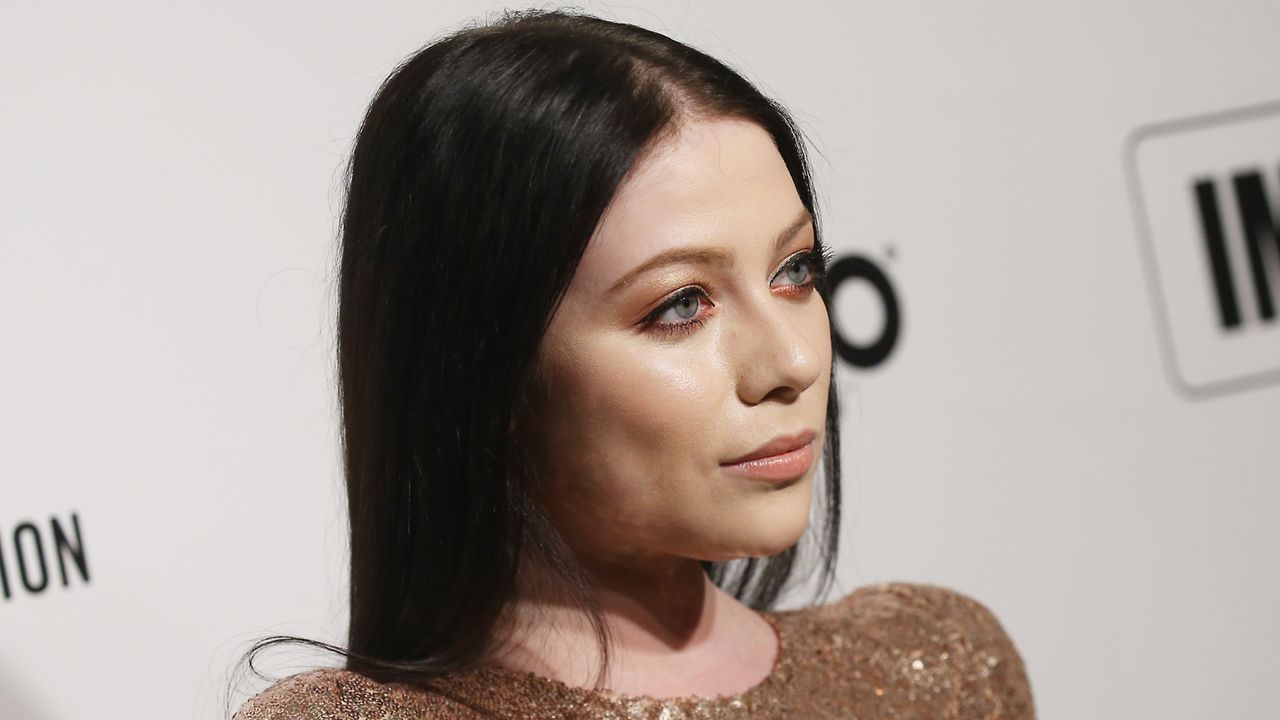 Michelle Trachtenberg wearing a rose gold sequined gown standing in front of a white step and repeat