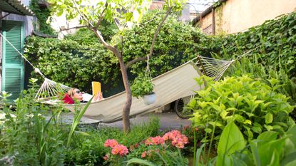 A woman lies in a hammock in her garden, reading a book. She looks relaxed.