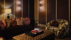 dark brown snug room with dark couch, patterned throw pillows, a tapestry print arm chair, and velvet upholstered ottoman