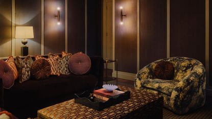 dark brown snug room with dark couch, patterned throw pillows, a tapestry print arm chair, and velvet upholstered ottoman