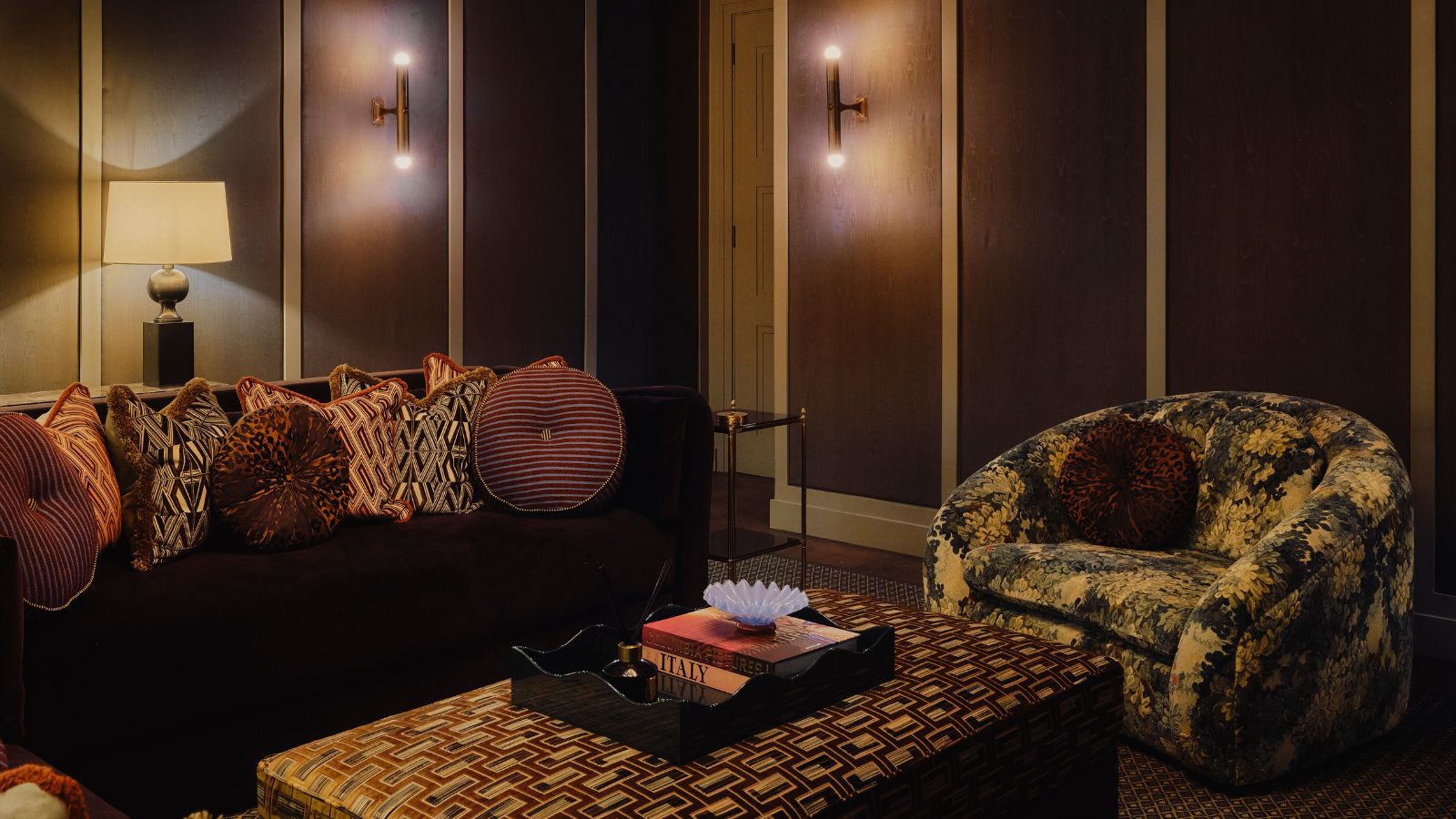 dark brown snug room with dark couch, patterned throw pillows, a tapestry print arm chair, and velvet upholstered ottoman