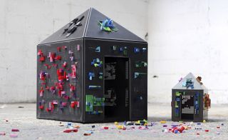 White rustic floor and walls, Dark grey and colourful Lego models of a large and a small house, colourful Lego pieces scattered on the floor around the houses
