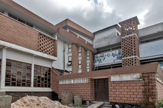 Daytime exterior image of Nwoko’s Oba Akenzua Cultural Centre in Benin City, brick walls, wooden window frames, sand mound and construction plank and concrete building, grey cloudy skyblocks,