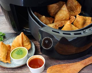 A black air fryer appliance with Asian samosas in basket and on plate adjacent to air fryer with assortment of chutneys, sauces and condiments