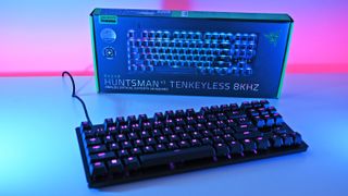 A Razer Huntsman V3 Tenkeyless keyboard with RGB lighting in front of its box. A vibrant pink and blue background creates an energetic, high-tech feel.