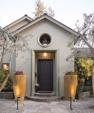 Exterior shot of front of gray house, stone path to front door, two large stone planters either side of small porch, light above black front door, small rounded window, trees and greenery surrounding the home