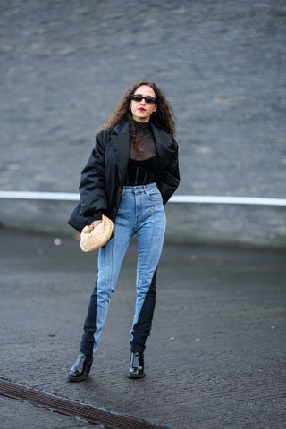A guest wears black sunglasses, a black turtleneck transparent tulle t-shirt, a black velvet corset, a black nylon puffer jacket, a beige braided shiny leather Jodie handbag from Bottega Veneta, blue and black denim bicolored embroidered seams pattern skinny denim pants from Mugler, black shiny leather varnished block heels ankle boots , outside A. Roege Hove, during the Copenhagen Fashion Week Autumn/Winter 2023 on February 01, 2023 in Copenhagen, Denma
