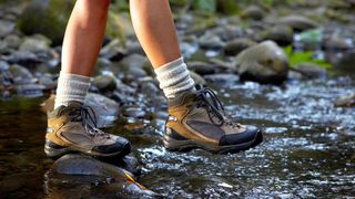 A hiker traverses a stream in brown hiking boots.