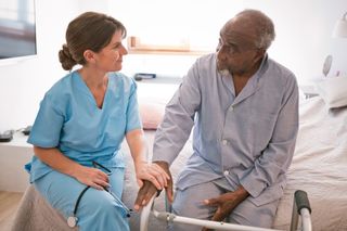 Female nurse talking with elderly man through glaucometer. They are sitting on bed.