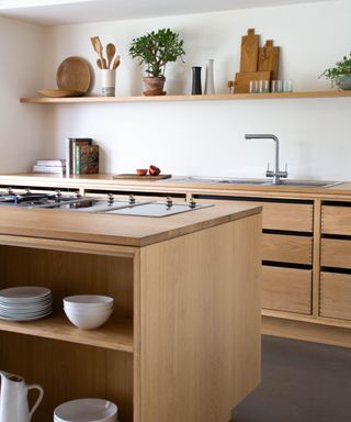A light wood kitchen island with open shelves with white plates and bowls, a silver hob on it, and wooden cabinets behind it with a silver faucet and books on it