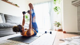 A woman exercises at home with a dumbbell on an exercise mat. She is lying down, feet and legs elevated upwards, holding the dumbbell with straight arms above her head. Behind her we see a couch and plants.