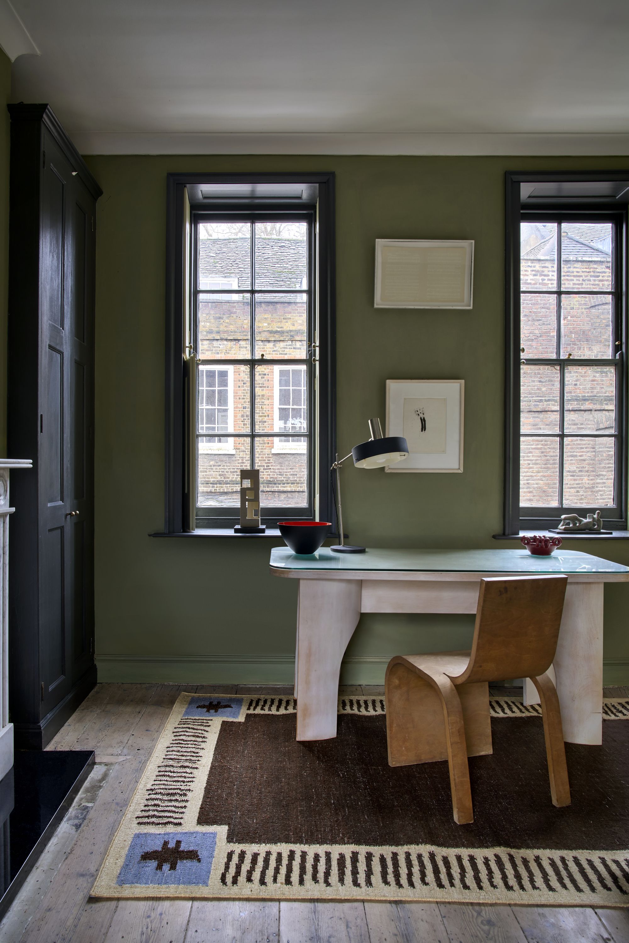 An olive green home office with black cabinetry and trim. There is a white desk with a wooden desk chair and a brown, blue, and cream rug underneath.