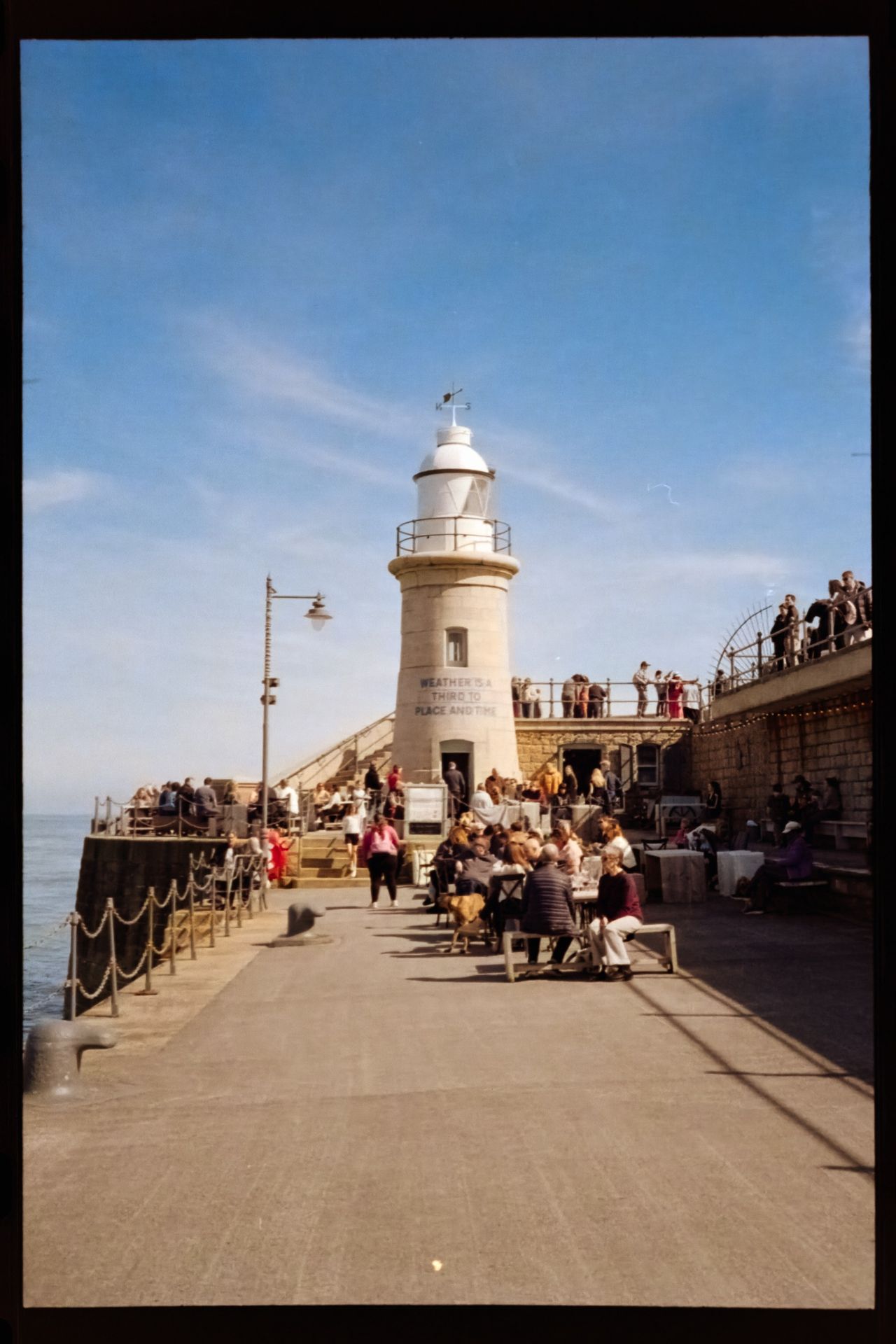 Cropped scan of a seaside pier centered on a small lighthouse, with people dining and walking around it under a clear blue sky.