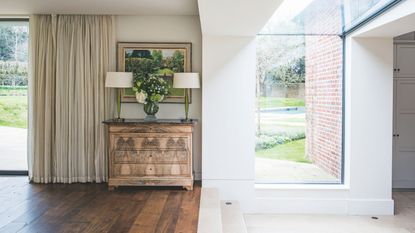 Bright transitional space with glass windows, wooden flooring, white walls, and a brown chest of drawers with two matching lamps with white shades, an artwork above, and cream curtains to the left.