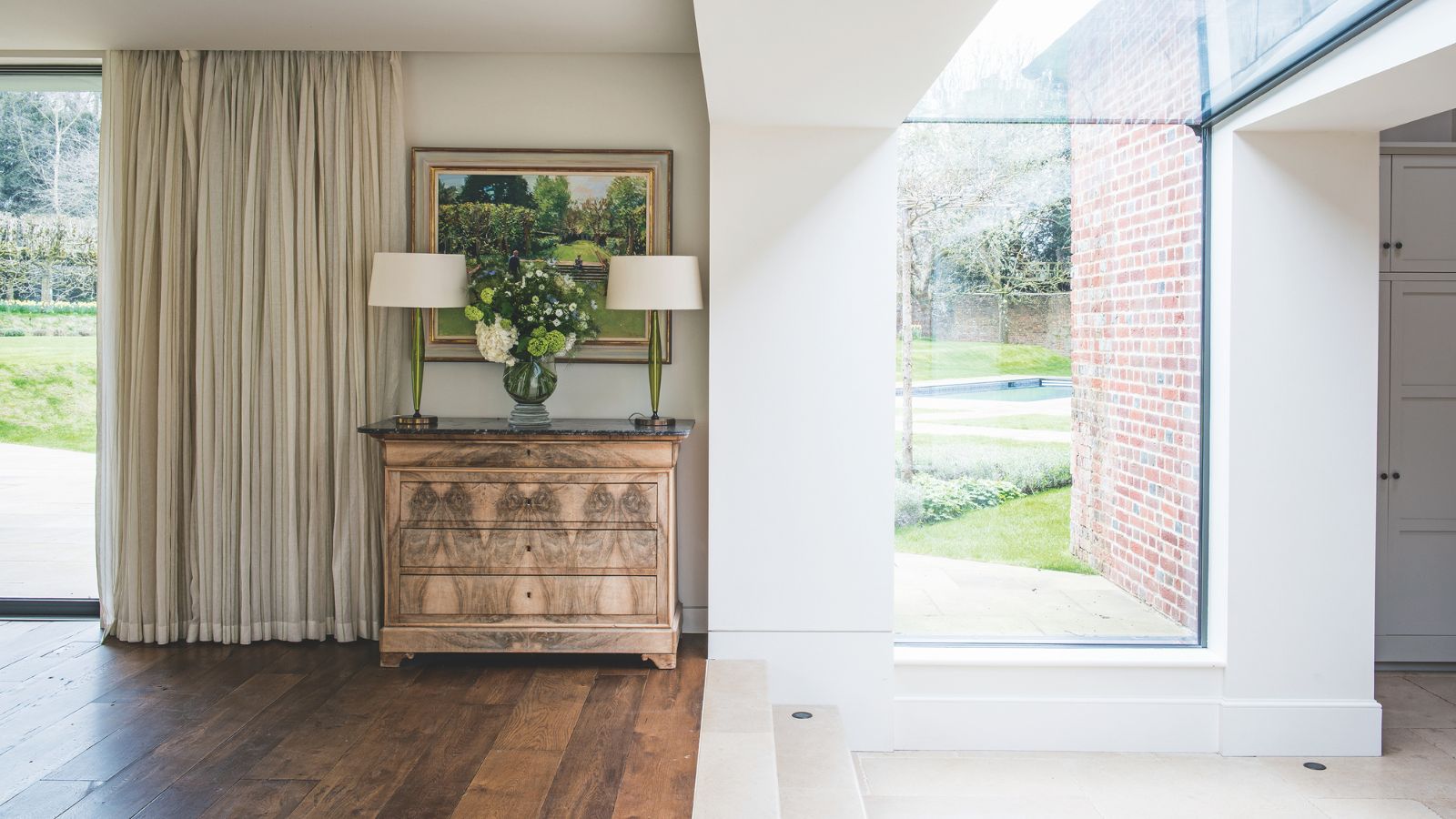 Bright transitional space with glass windows, wooden flooring, white walls, and a brown chest of drawers with two matching lamps with white shades, an artwork above, and cream curtains to the left.
