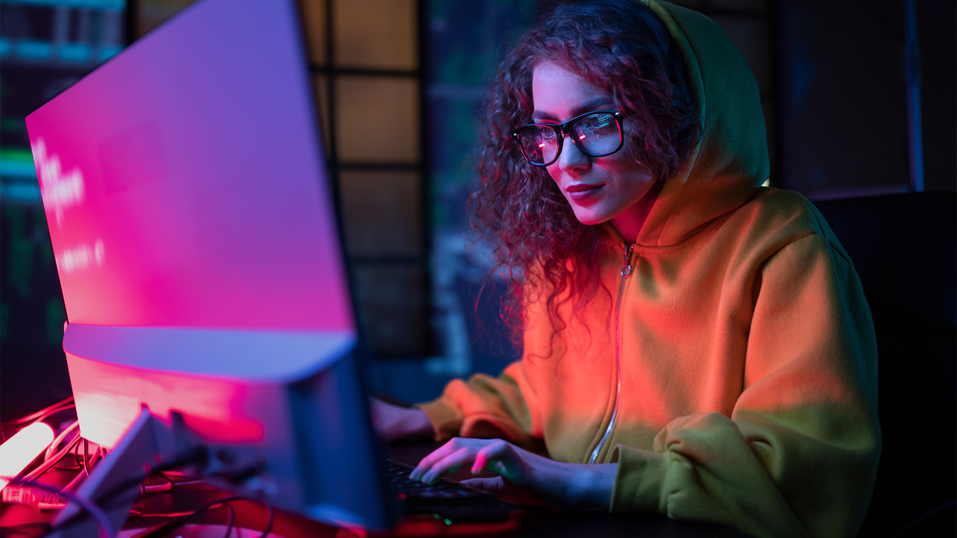 Female software developer wearing hoodie coding on a desktop computer in a dimly lit room with warm pink glowing light reflecting off computer monitor.