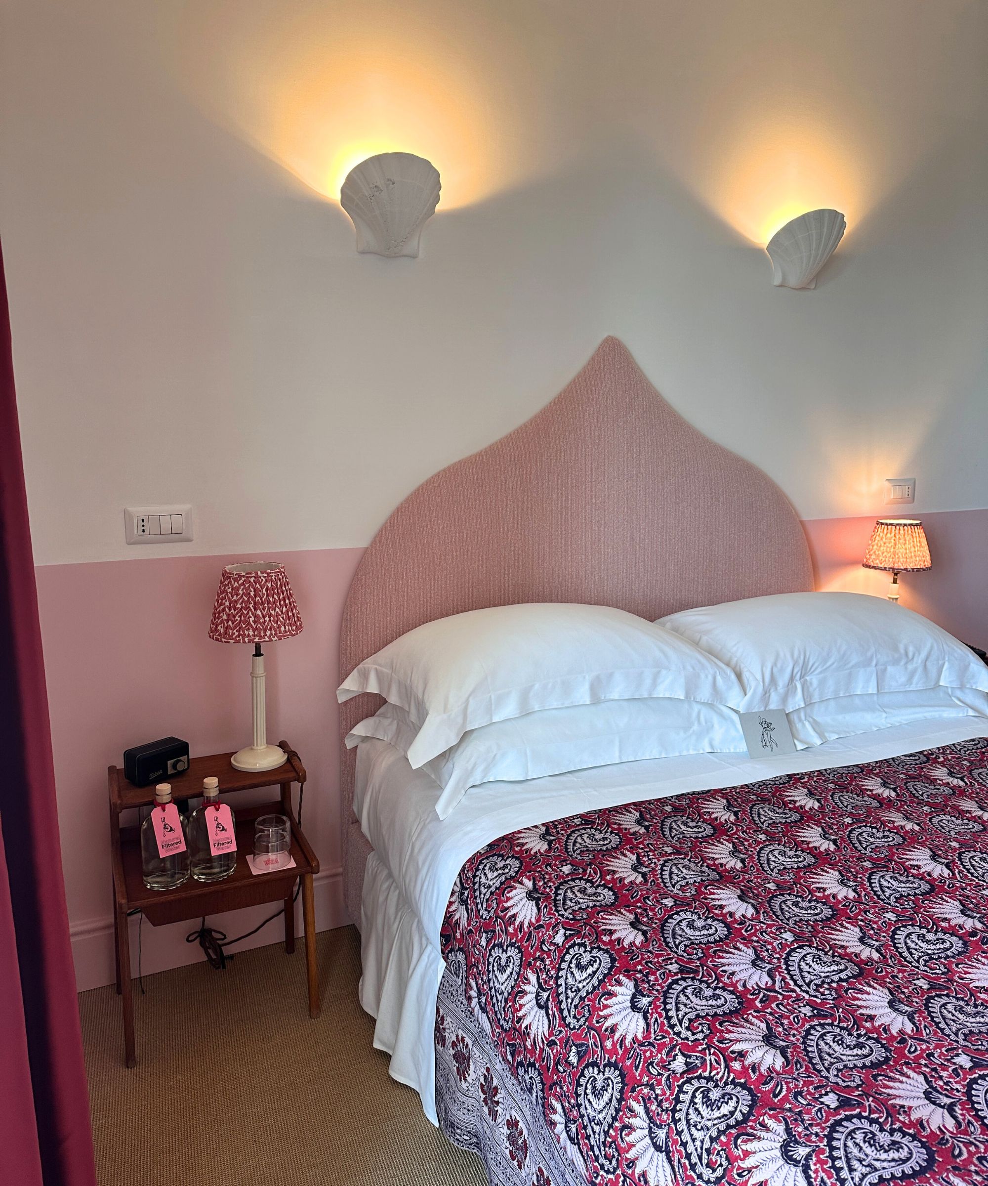 pink hotel bedroom with half painted pink and white walls, white shell wall sconce lights, a pale pink shaped headboard, white bed sheets and a block printed floral bed spread