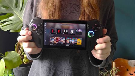 Hands holding ROG Xbox Ally X handheld with plants and green wall in backdrop.