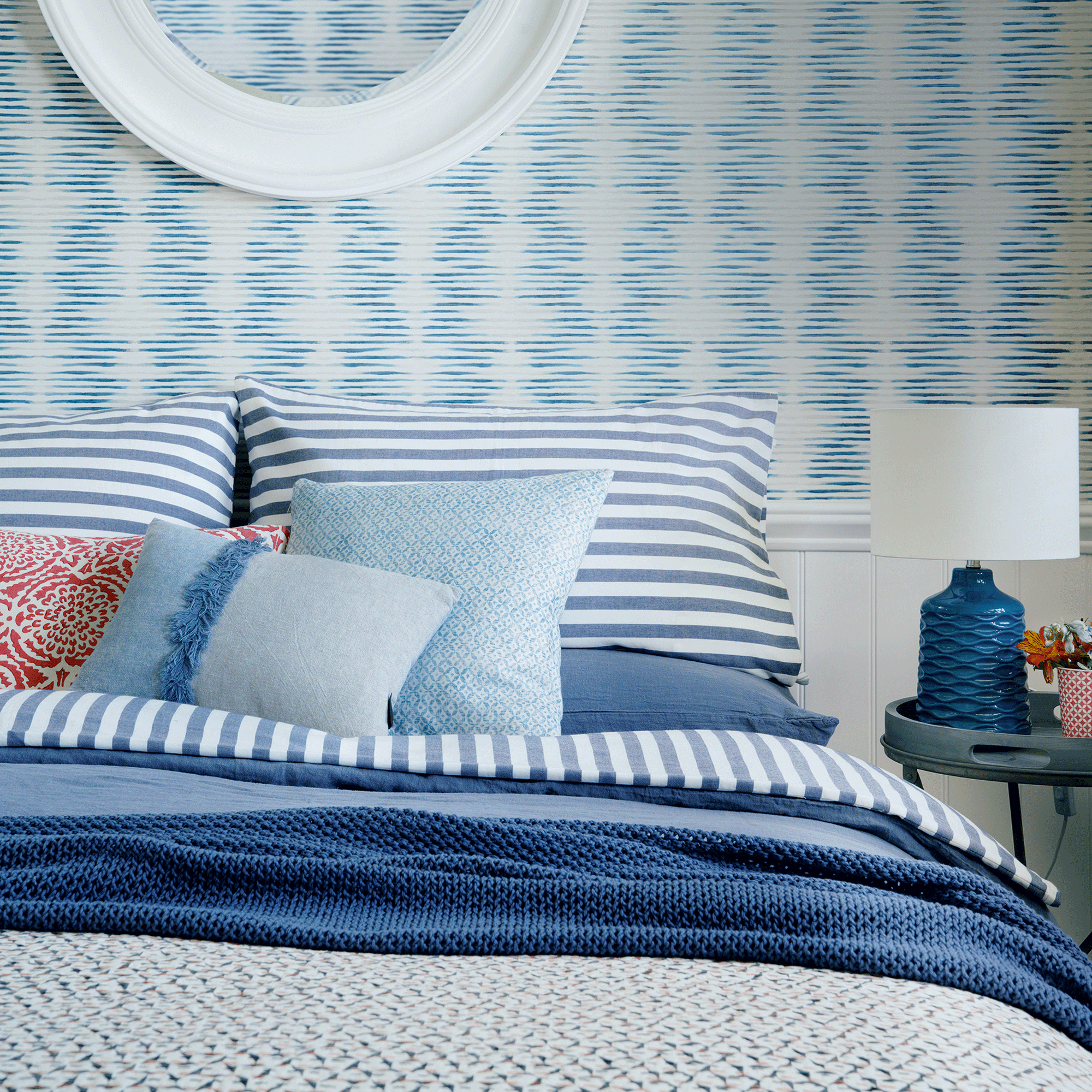 a blue and white bedroom with patterned wallpaper, striped bedding, and a selection of blue, white and red cushions