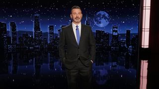 Jimmy Kimmel stands in front of his cityscape set with his hands in his pockets. He is wearing a blue tie and black suit.