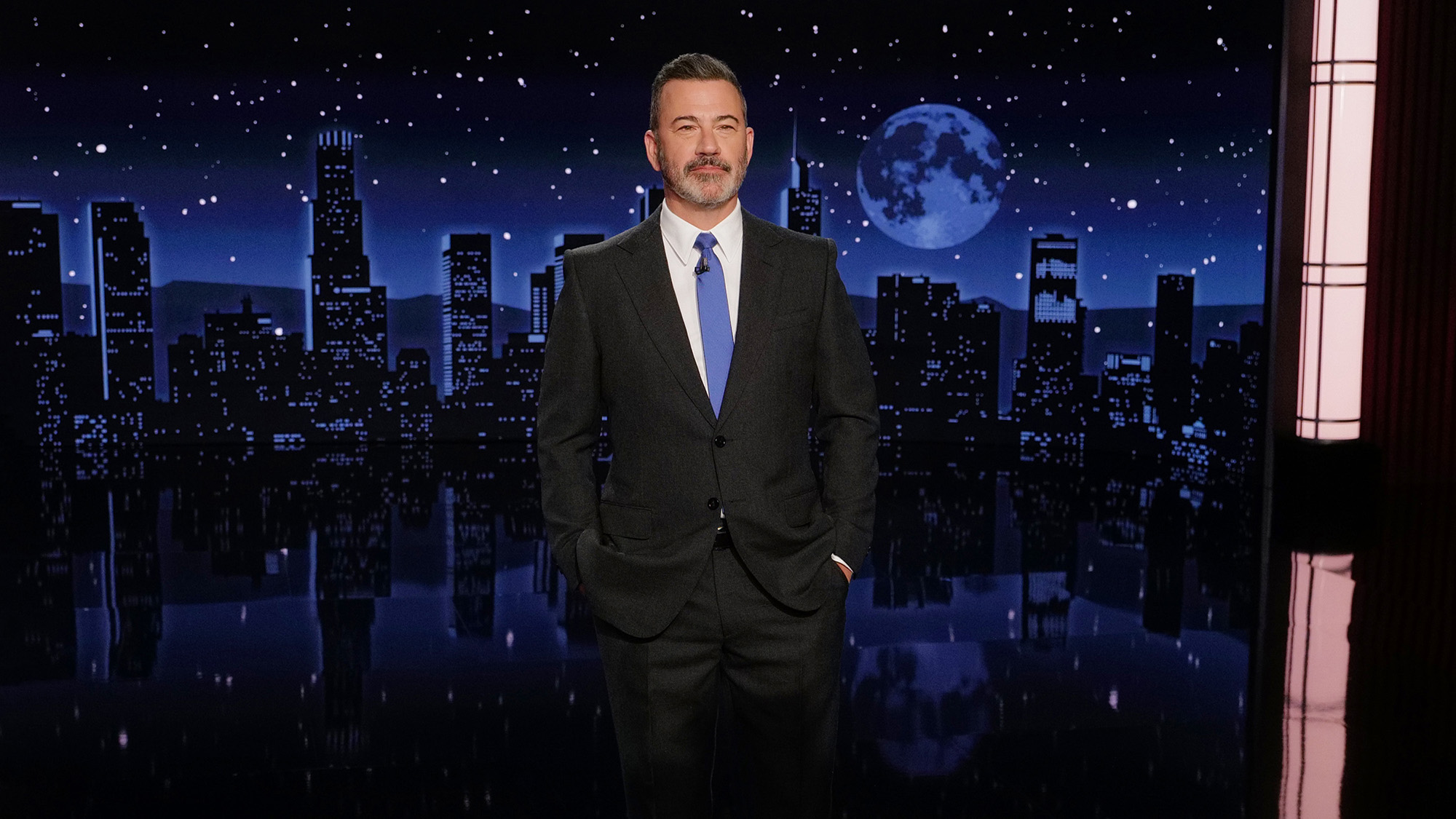 Jimmy Kimmel stands in front of his cityscape set with his hands in his pockets. He is wearing a blue tie and black suit.