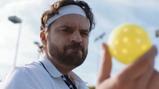 Jake Johnson as Dusty Boyd is shown in The Dink, looking at a pickleball with disdain.