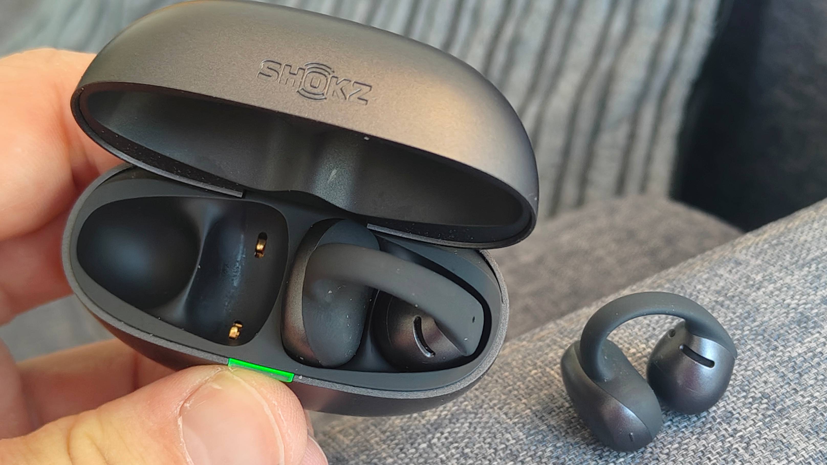 The Shokz OpenDots Air case, with a single bud inside, and the other on a nearby sofa arm.