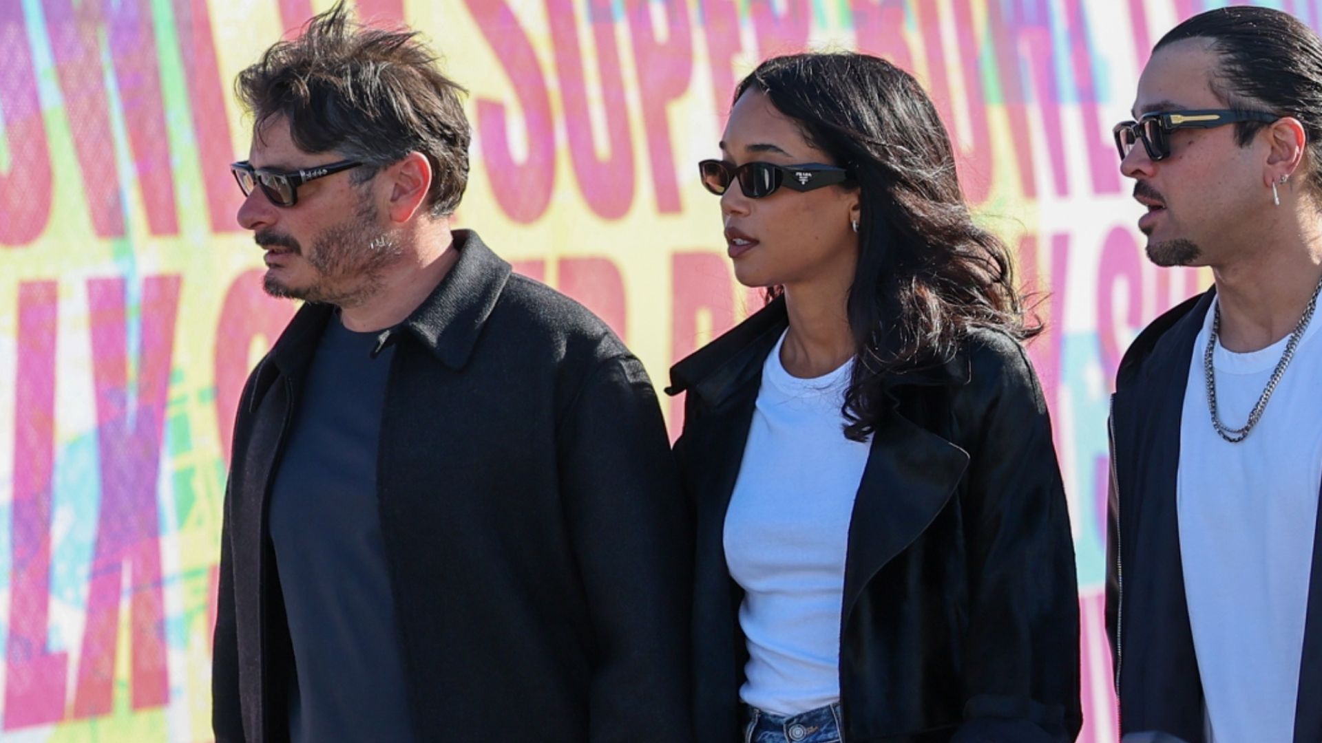 Laura Harrier walks outside wearing high-waisted straight-leg jeans with heeled boots, a white tee and a black trench coat.