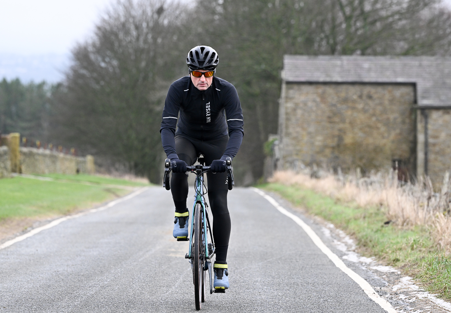 Man riding directly at the camera on a green Cervelo road bike wearing black tights, black jacket and blue shoes, up a country lane.