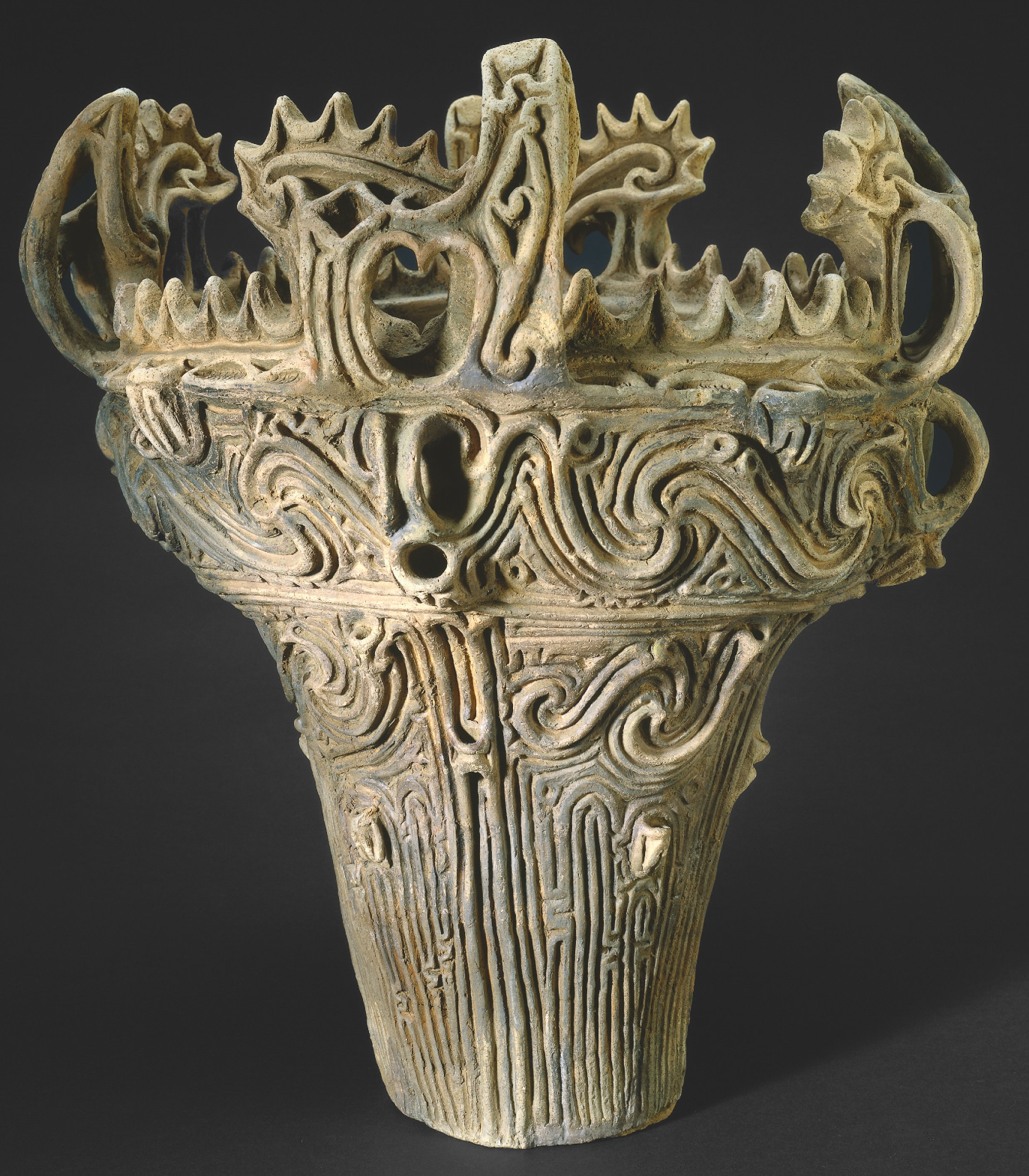 a light clay vessel made from coils that tapers from the top to the bottom and looks like it has stylized flames at the rim