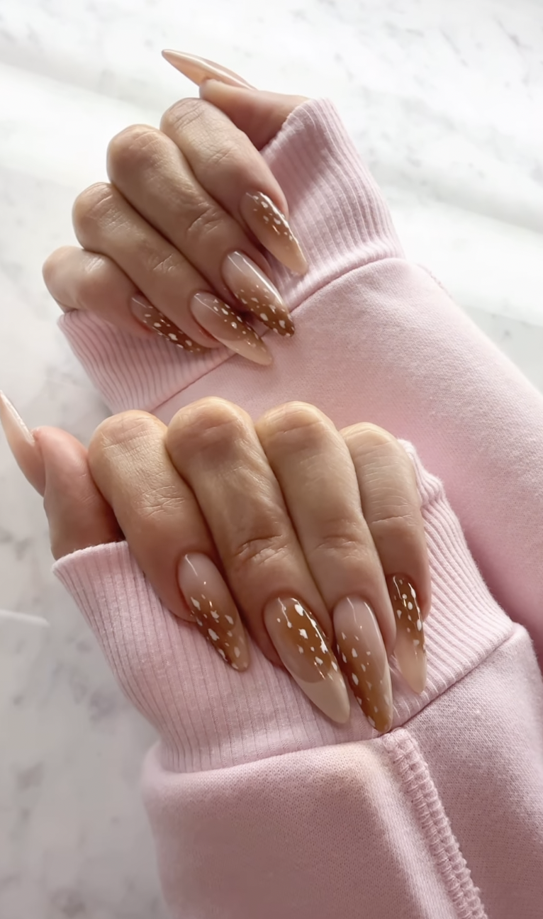 Long almond nails with warm brown tips and little white deer spots, hands in pink sweater sleeves
