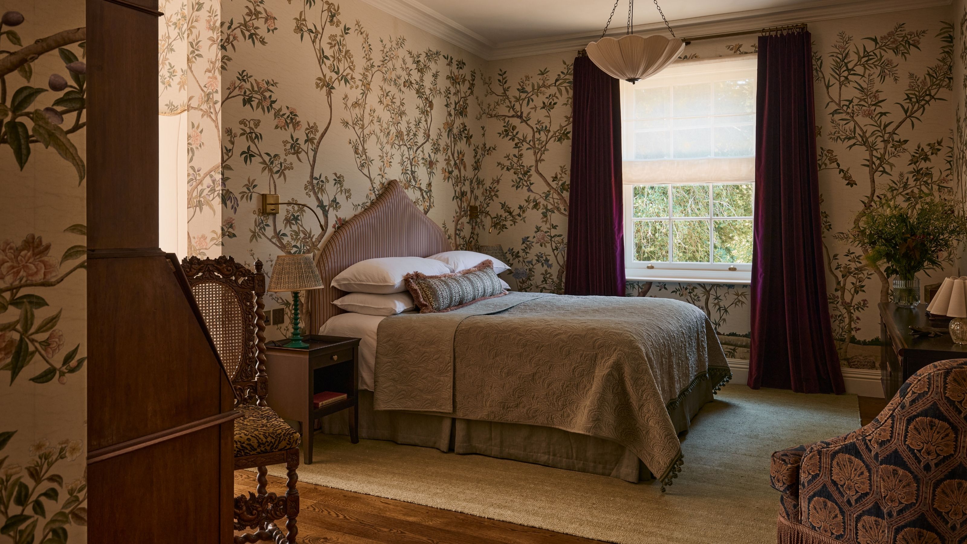 a wallpaper wrapped guest room with dark red draped, striped headboard and antique furniture