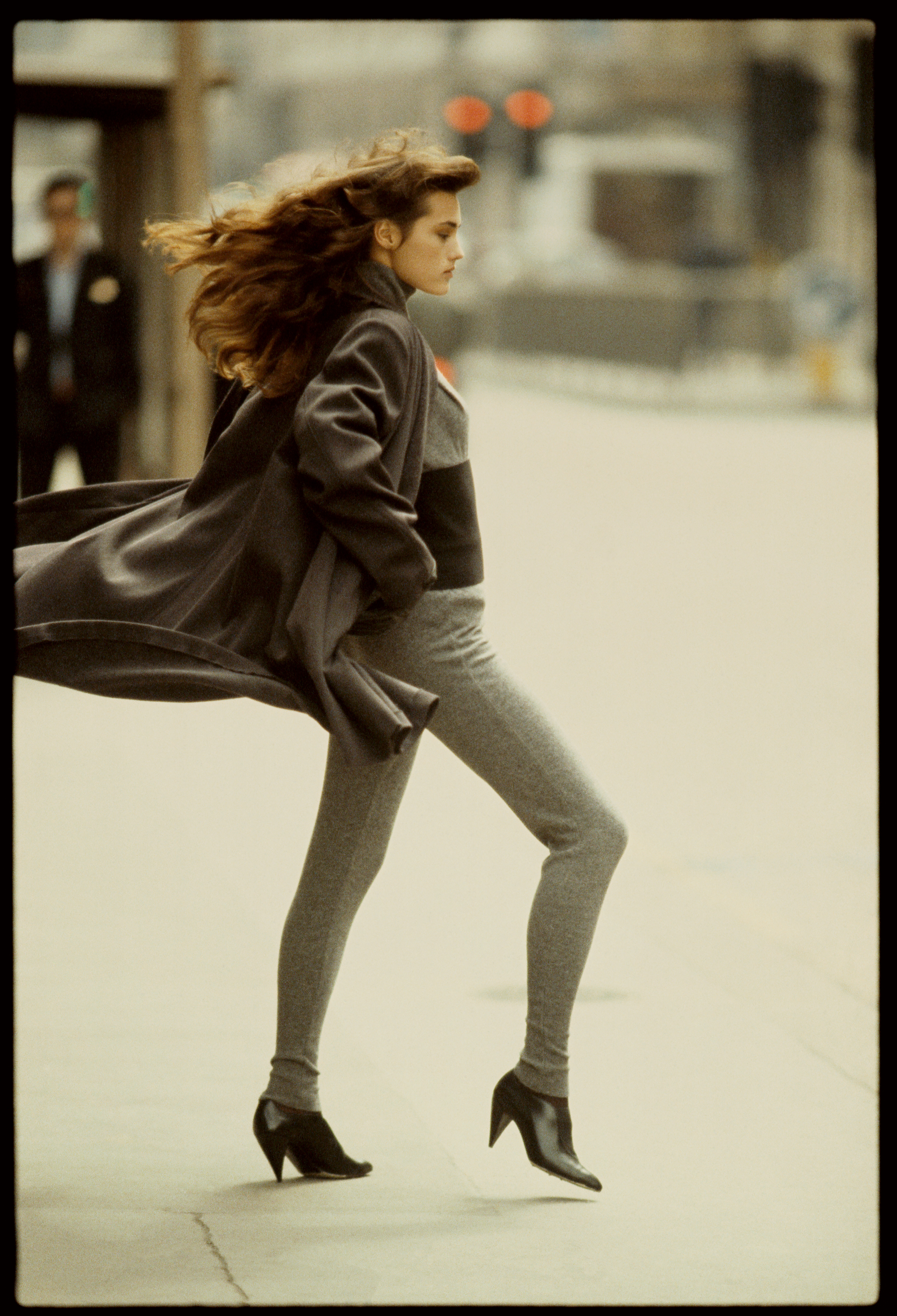 A model in a grey jumper and trousers strides forward mid-step, her hair and coat caught streaming in the wind