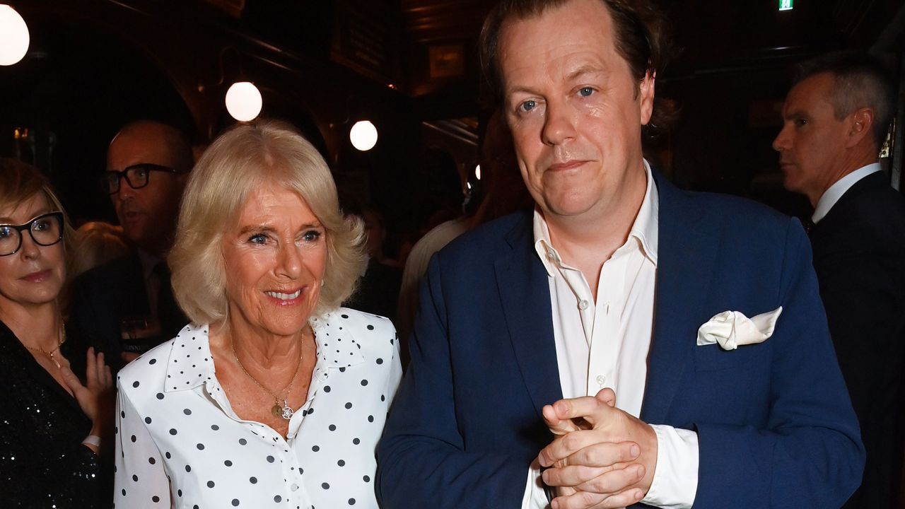 Queen Camilla wearing a white polka dot dress smiling and posing with her son Tom Parker Bowles who is wearing a nvay suit and white shirt