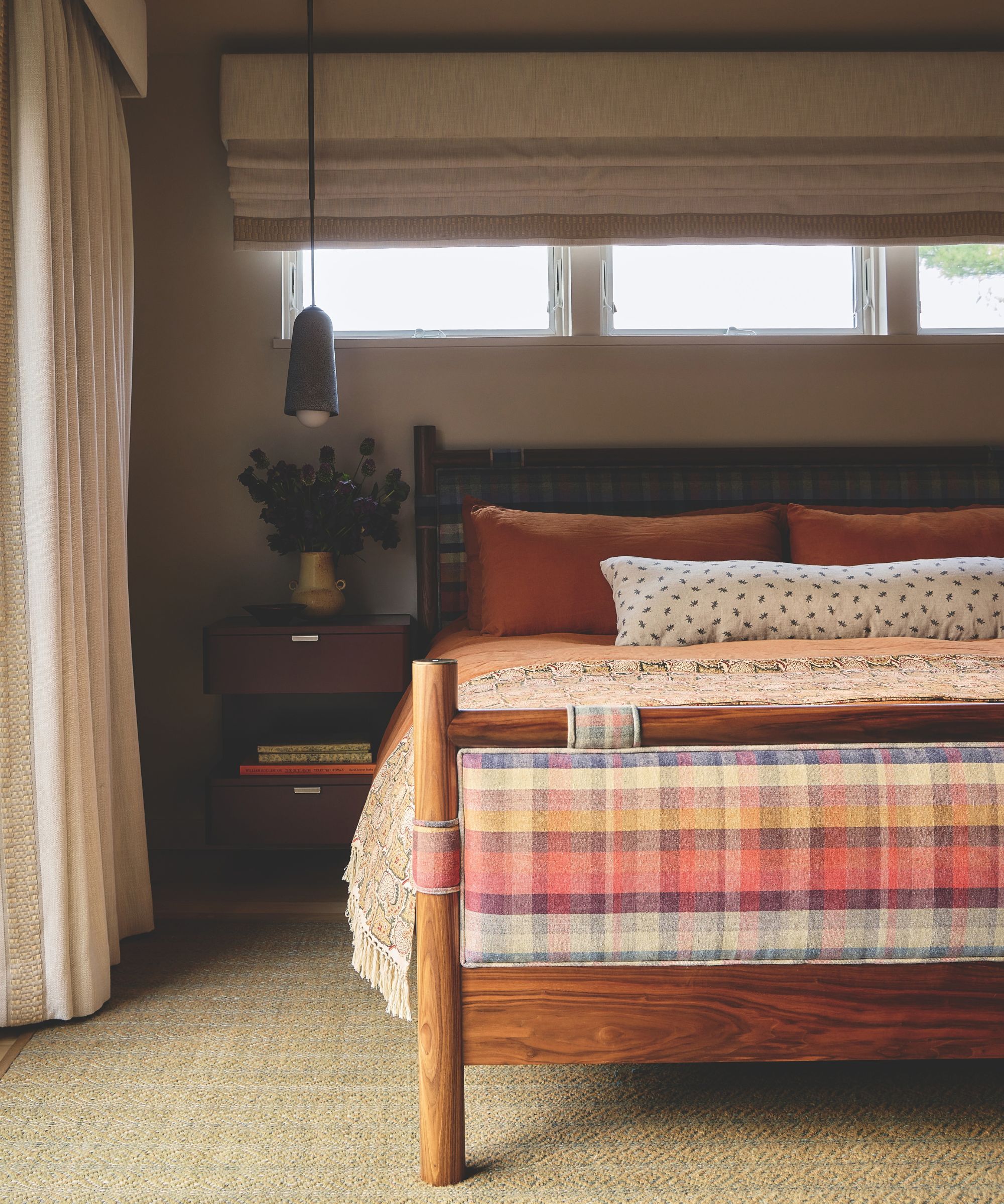Cozy bedroom setting with a wooden bed frame, colorful plaid fabric footboard, terracotta pillows, and low pendant lighting.