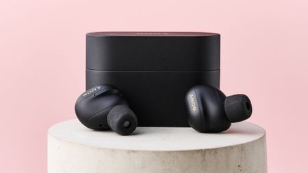 The Sony WF-1000XM6 lying in front of their case on top of a stone pillar in front of a pink background.