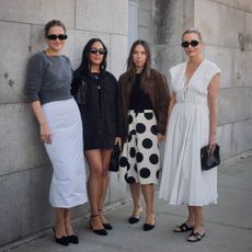 Four women pose for a picture. Eliza wears gray sweater and white pencil skirt. Nicole wears a black dress. Lauren wears a polka dot skirt, black top, and brown bomber. Kristen wears white dress.