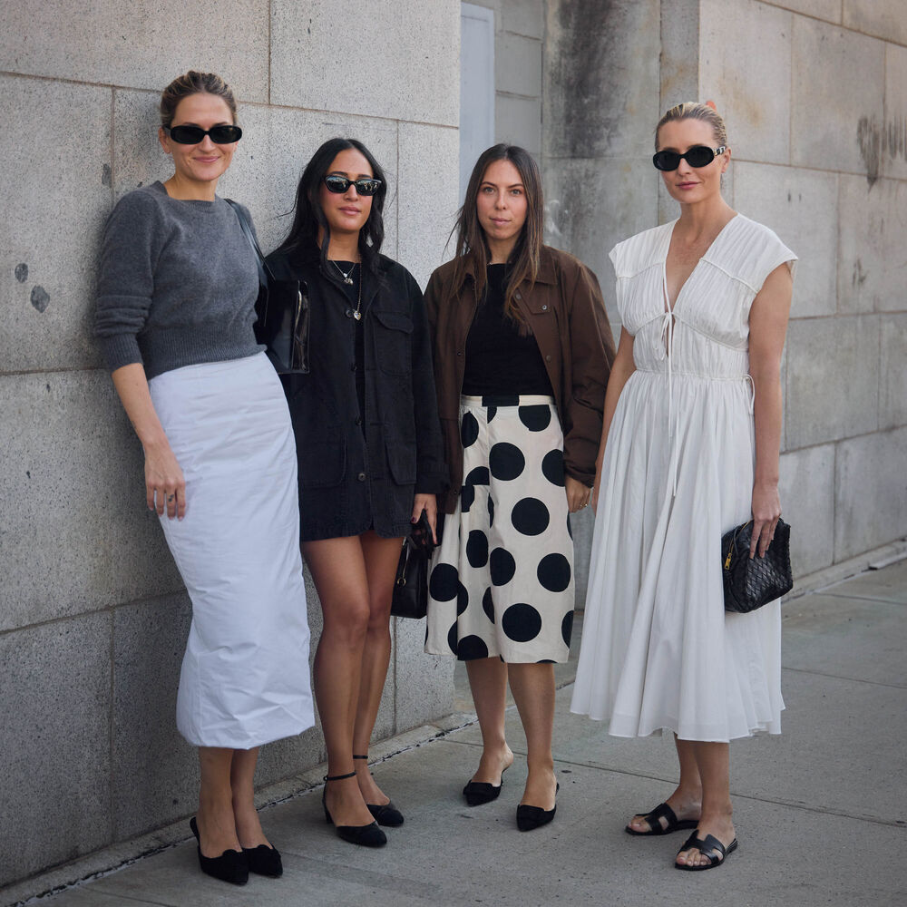Four women pose for a picture. Eliza wears gray sweater and white pencil skirt. Nicole wears a black dress. Lauren wears a polka dot skirt, black top, and brown bomber. Kristen wears white dress.