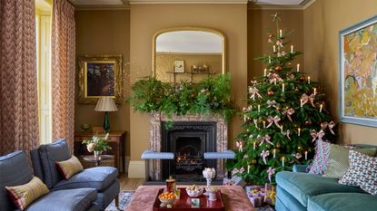 Cozy, traditional living room with mustard-yellow/brown walls. A large Christmas tree decorated with pink bows stands next to a fireplace mantle adorned with greenery. The seating includes a navy-blue sofa and a teal armchair.