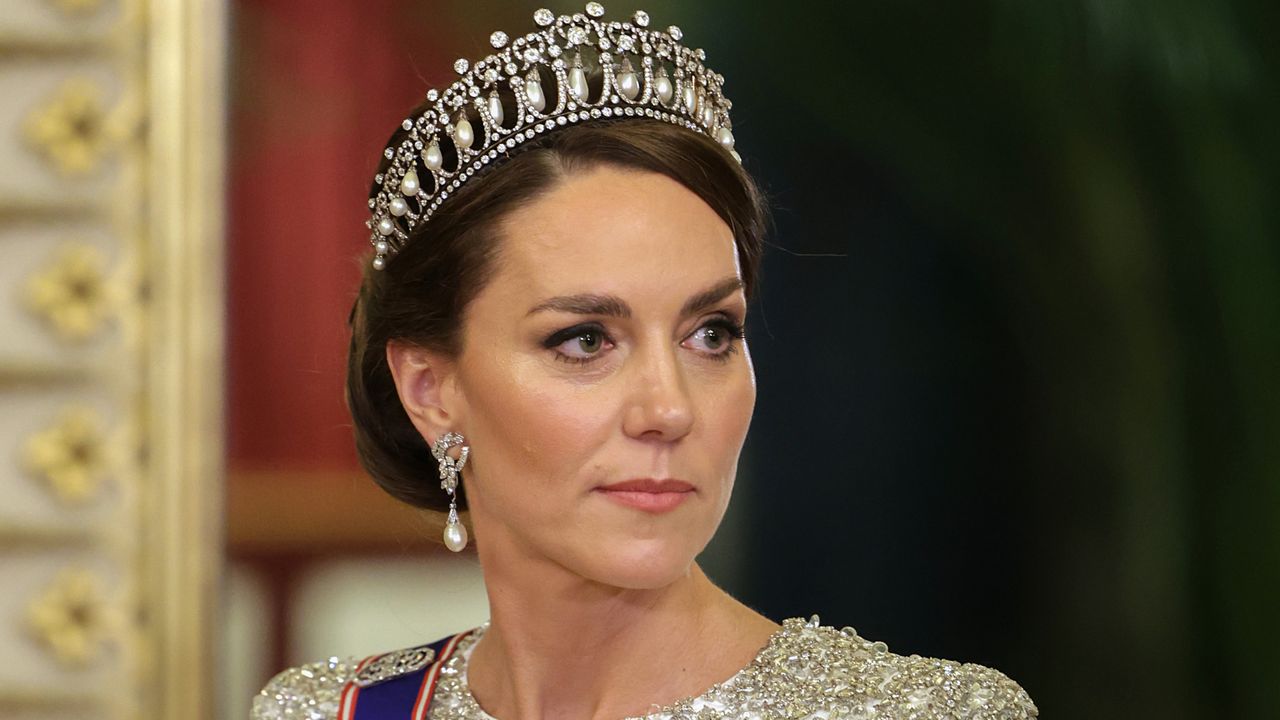 A shoulders-up photo of Kate Middleton wearing the Lover&#039;s Knot tiara and a white embellished gown with pearl earrings looking to the side with a serious expression