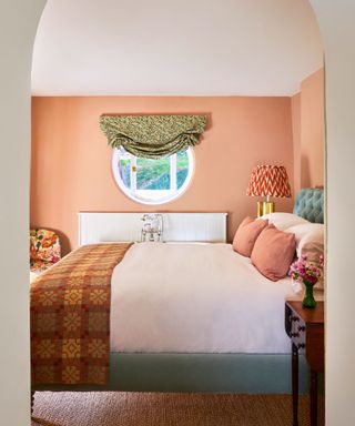 Archway into bedroom with peach walls, round window with pleated green floral treatment, cozy double bed with teal frame, patterned throw, arm chair and lamp