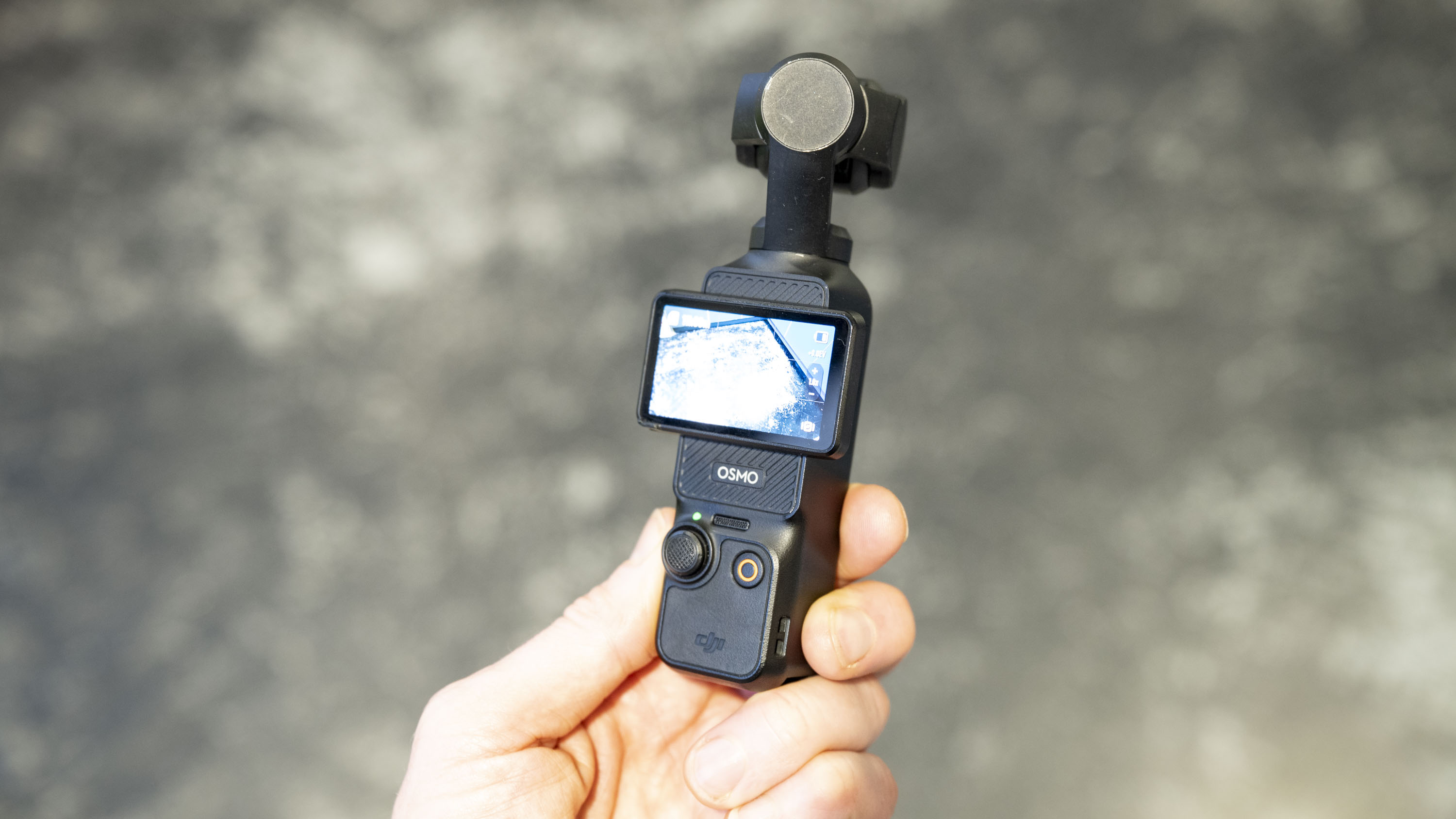 DJI Osmo Pocket 3 vlogging camera in user's hand, grey floor in background