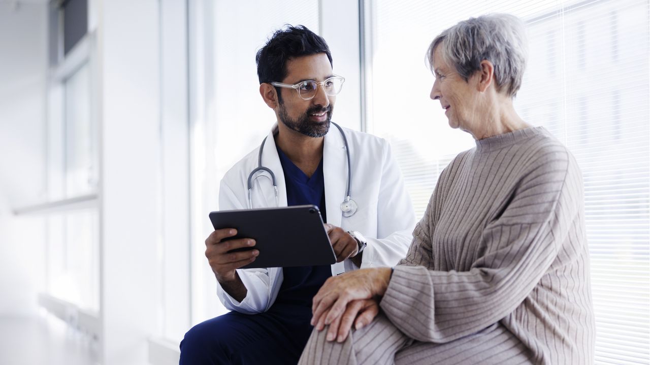 A doctor with a clipboard sits and talks with an older woman in a hospital corridor.