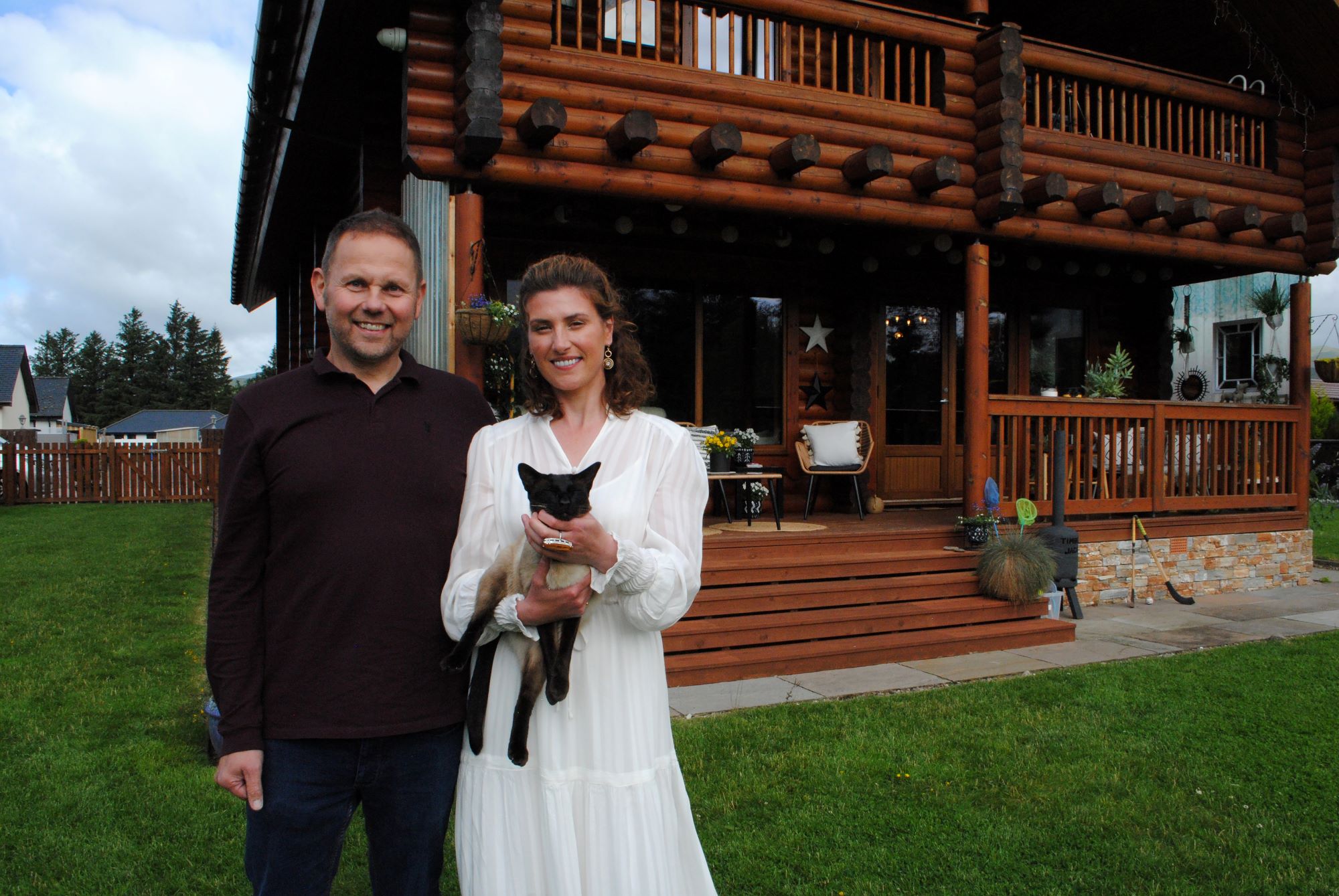 Jack and Penny standing in front of their log cabin home with their pet dog