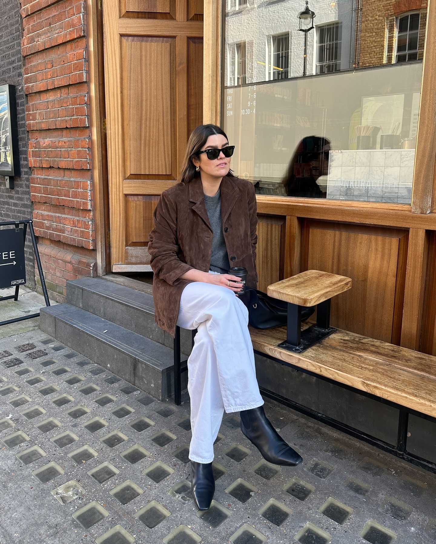 Influencer @theannaedit sits on a bench outside wearing a brown suede blazer with a grey jumper, white jeans and black pointed-toe boots.