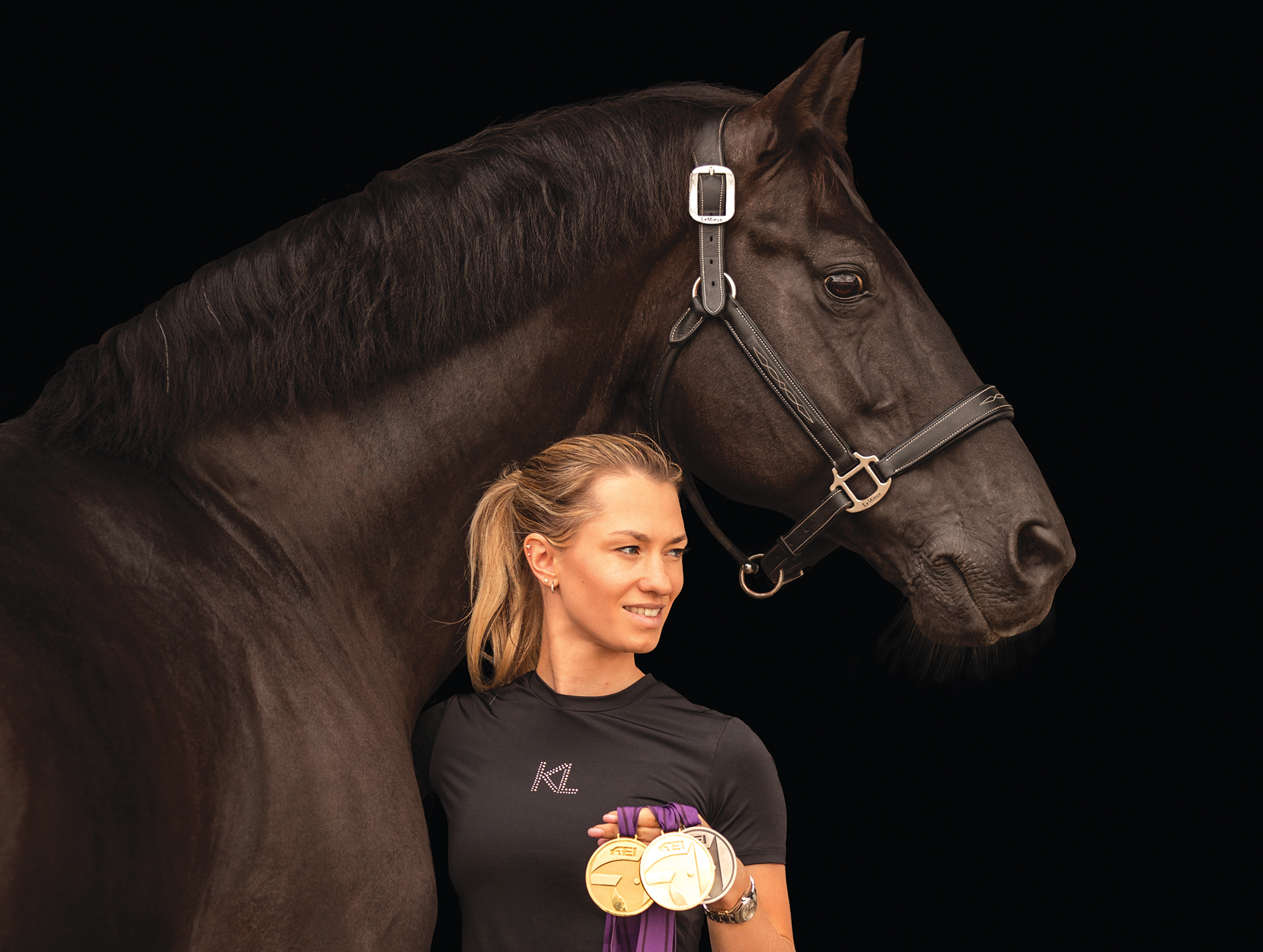 Lottie Fry holds her world championship medals as she stands beside a relaxed looking Galmourdale set against a black background
