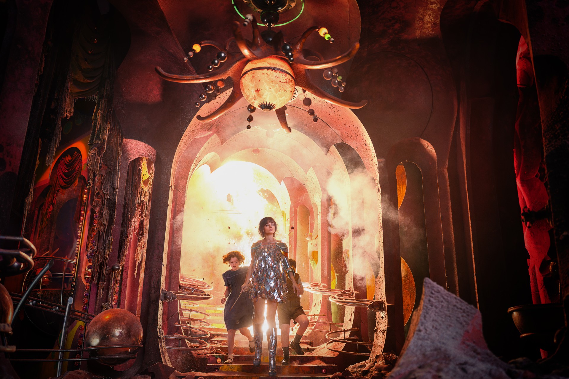 Jadis, in a mirrored minidress, shoulder armour and silver knee boots, strides down steps out of an explosion in a crumbling, rose-red palace, with Polly and Digory behind her