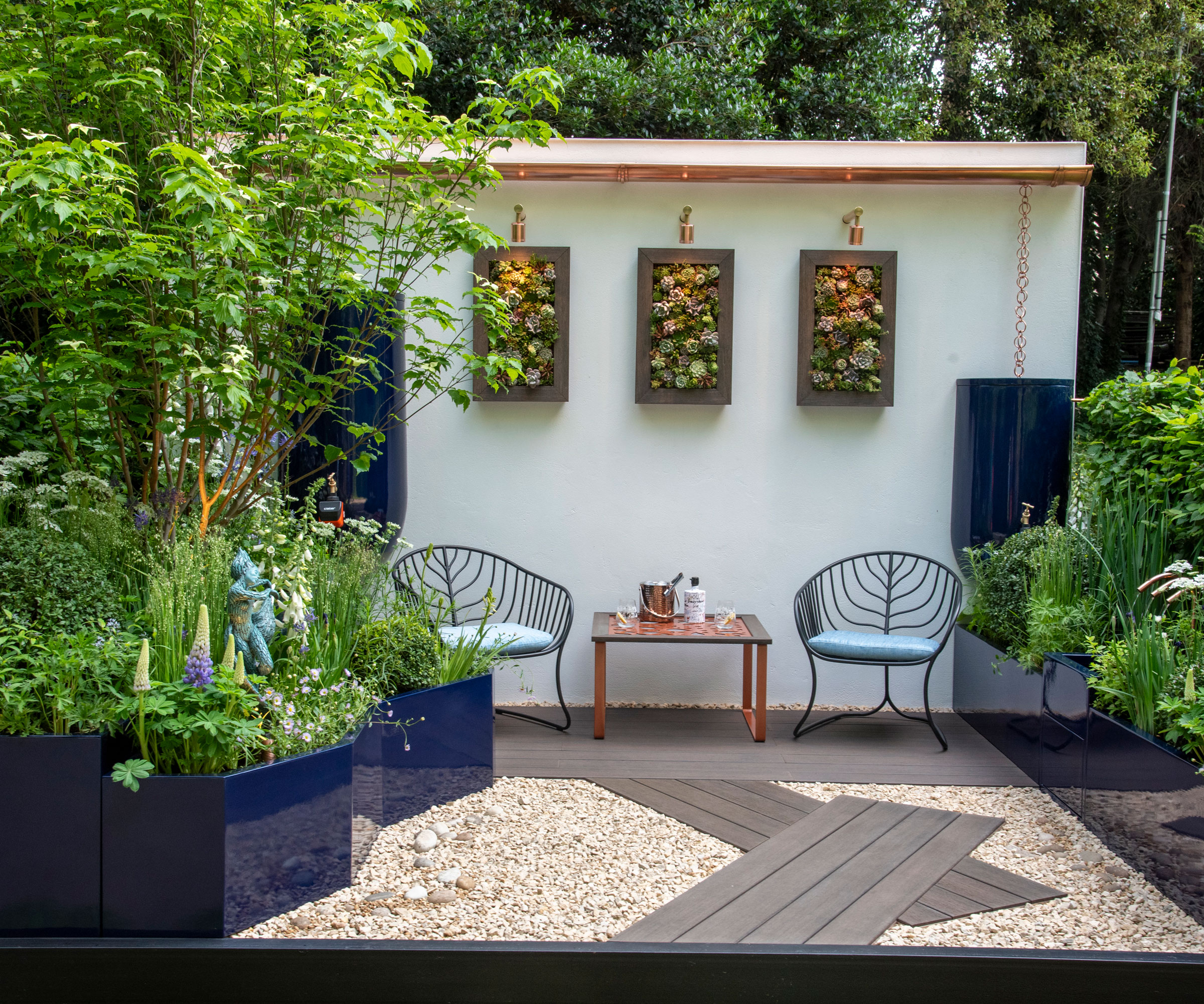 small courtyard garden design with garden furniture, decking and gravel area, raised beds with colourful planting, rain chain with wall-hung water storage, small trees and garden art hanging on a whitewashed wall