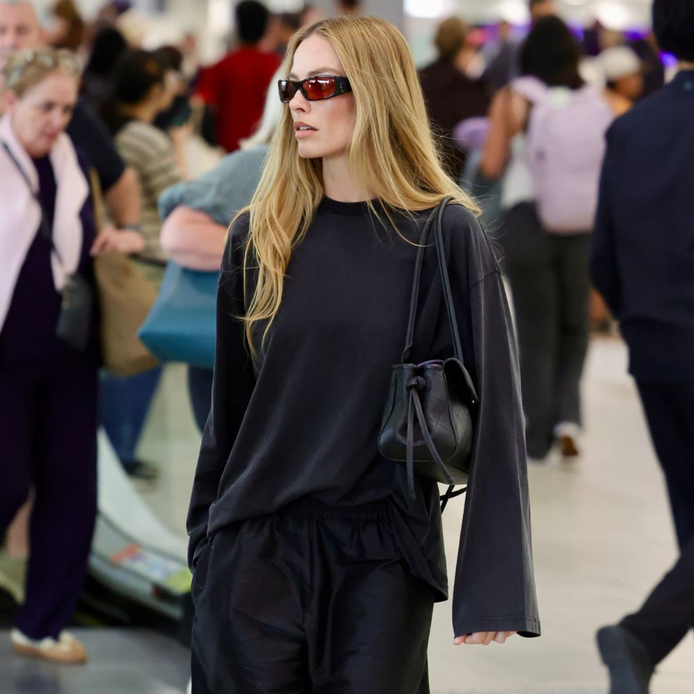 Margot Robbie walks through the airport wearing mesh ballet flats with long shorts and a black long-sleeve t-shirt.