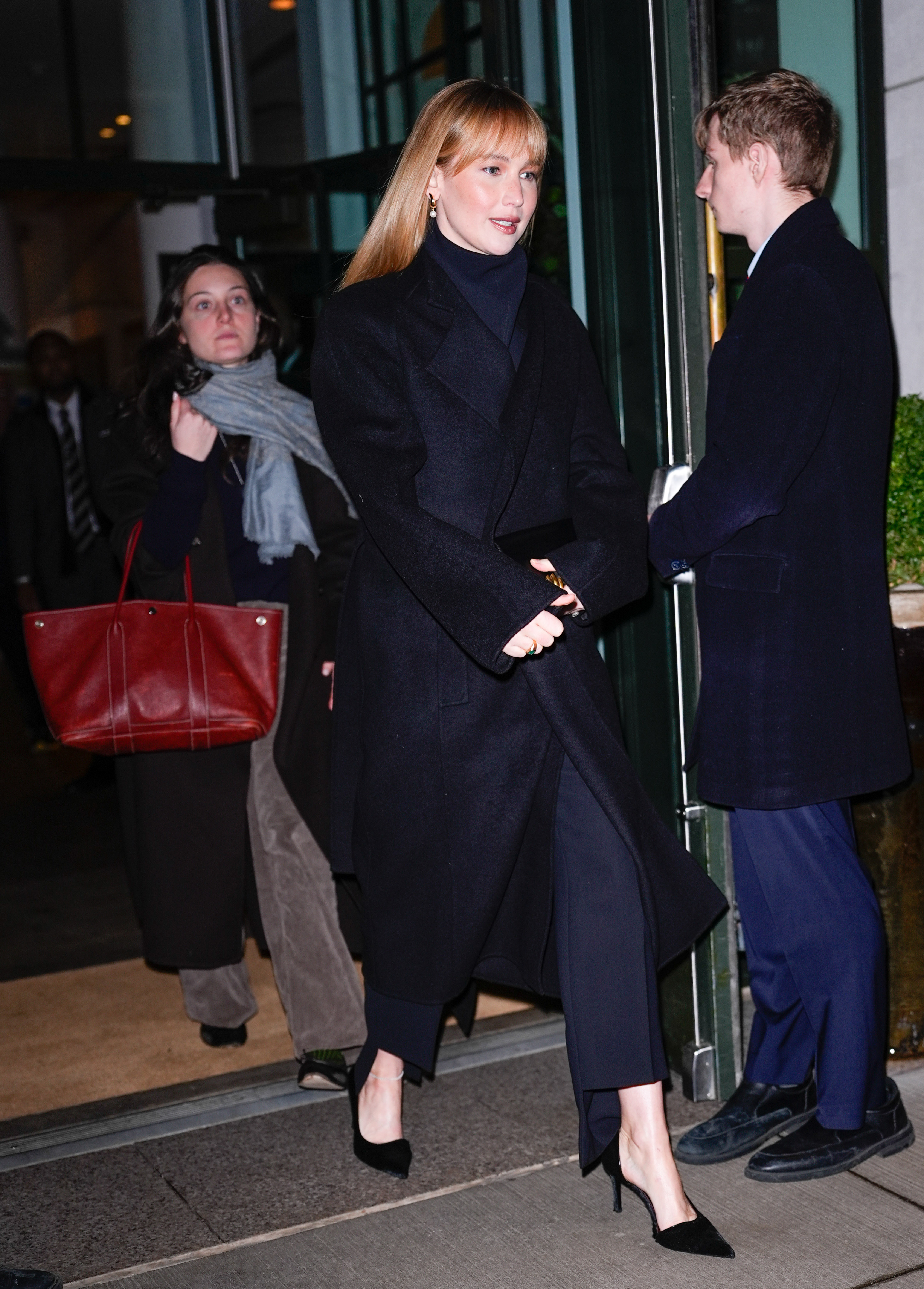 Jennifer Lawrence steps outside wearing a black coat with navy cigarette trousers with a black coat and black asymmetric heels.